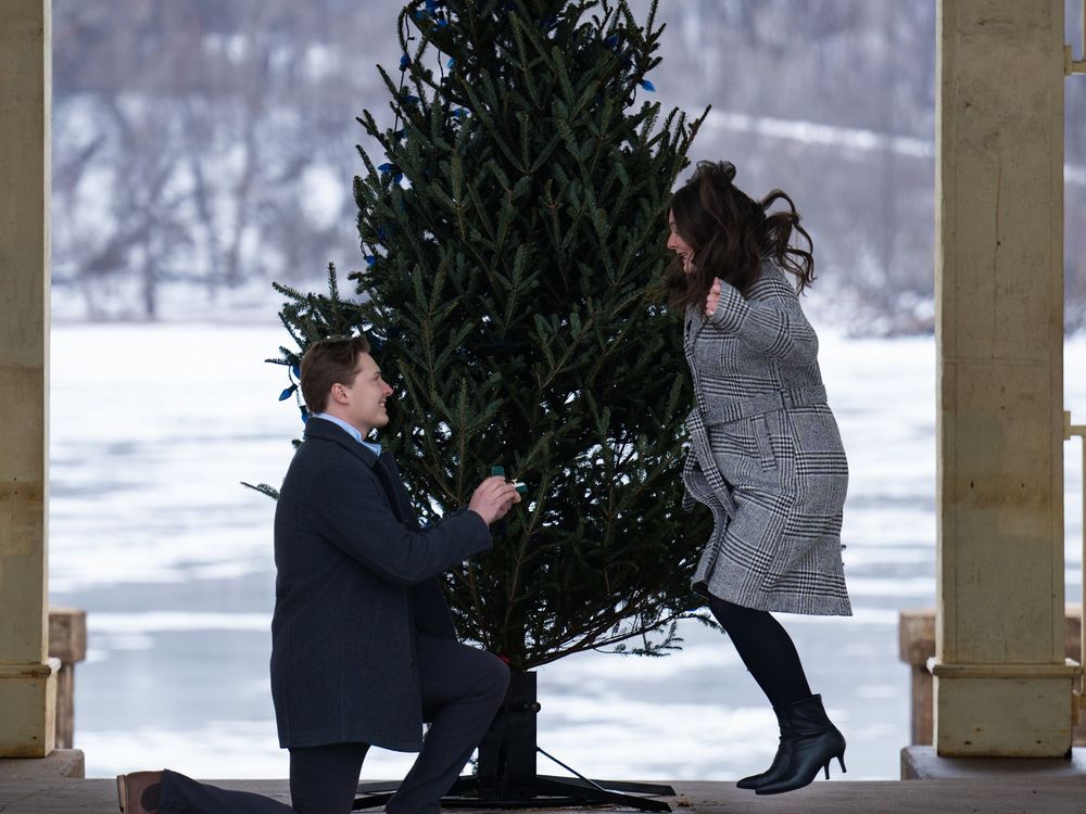A man kneels with a ring box as a woman leaps in surprise beside a winter pine