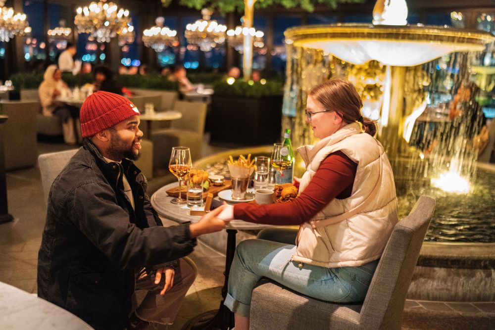 A man in a red beanie kneels beside a woman's chair in a chandelier-lit restaurant