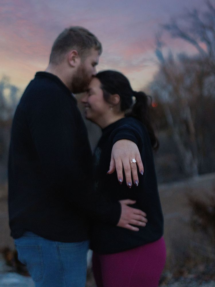 A newly engaged couple embraces at dusk as the woman shows her ring