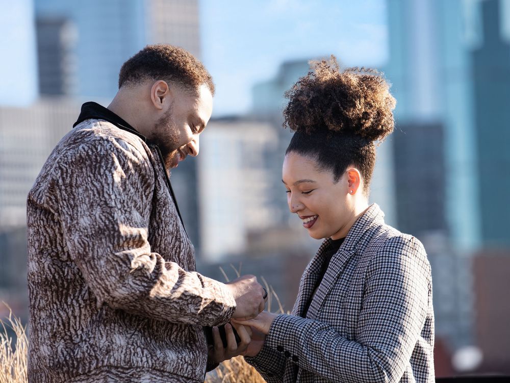 A man slides an engagement ring onto a smiling woman's finger with city towers behind