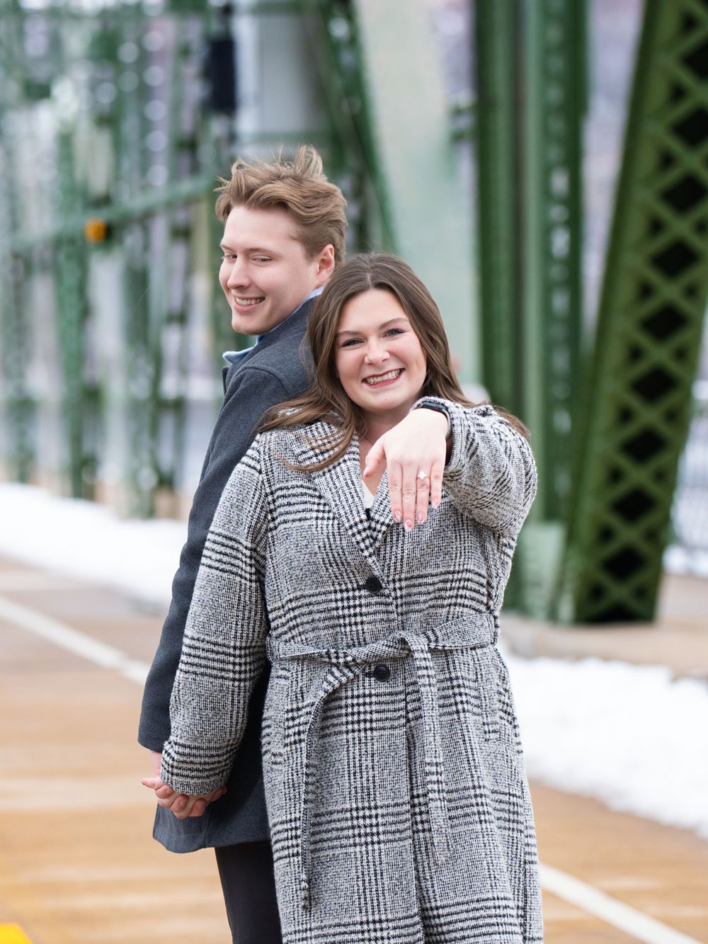 A smiling woman shows her engagement ring while her partner stands behind her on a steel truss bridge