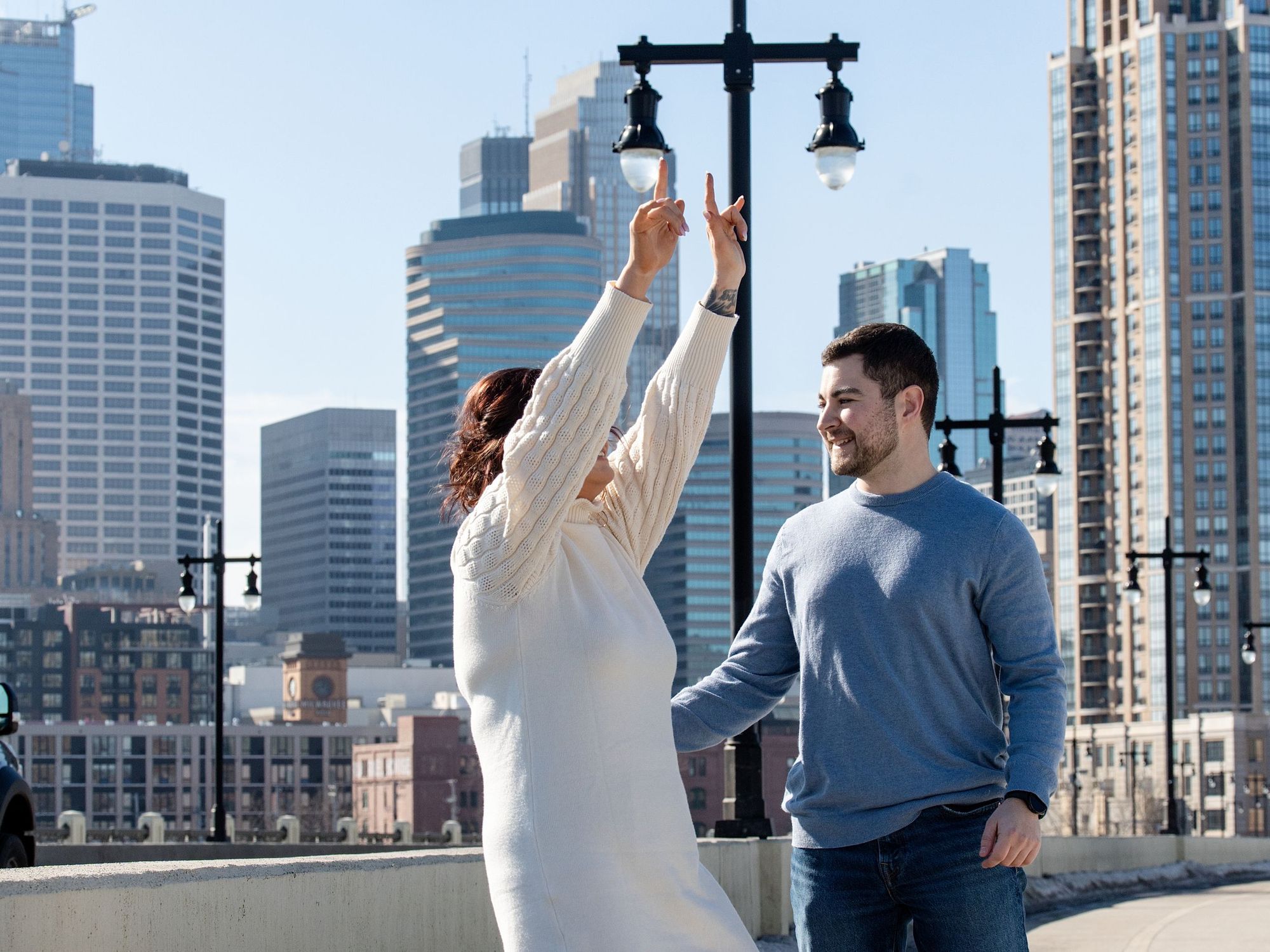 A newly engaged woman throws her arms up in celebration beside her partner with a downtown skyline behind