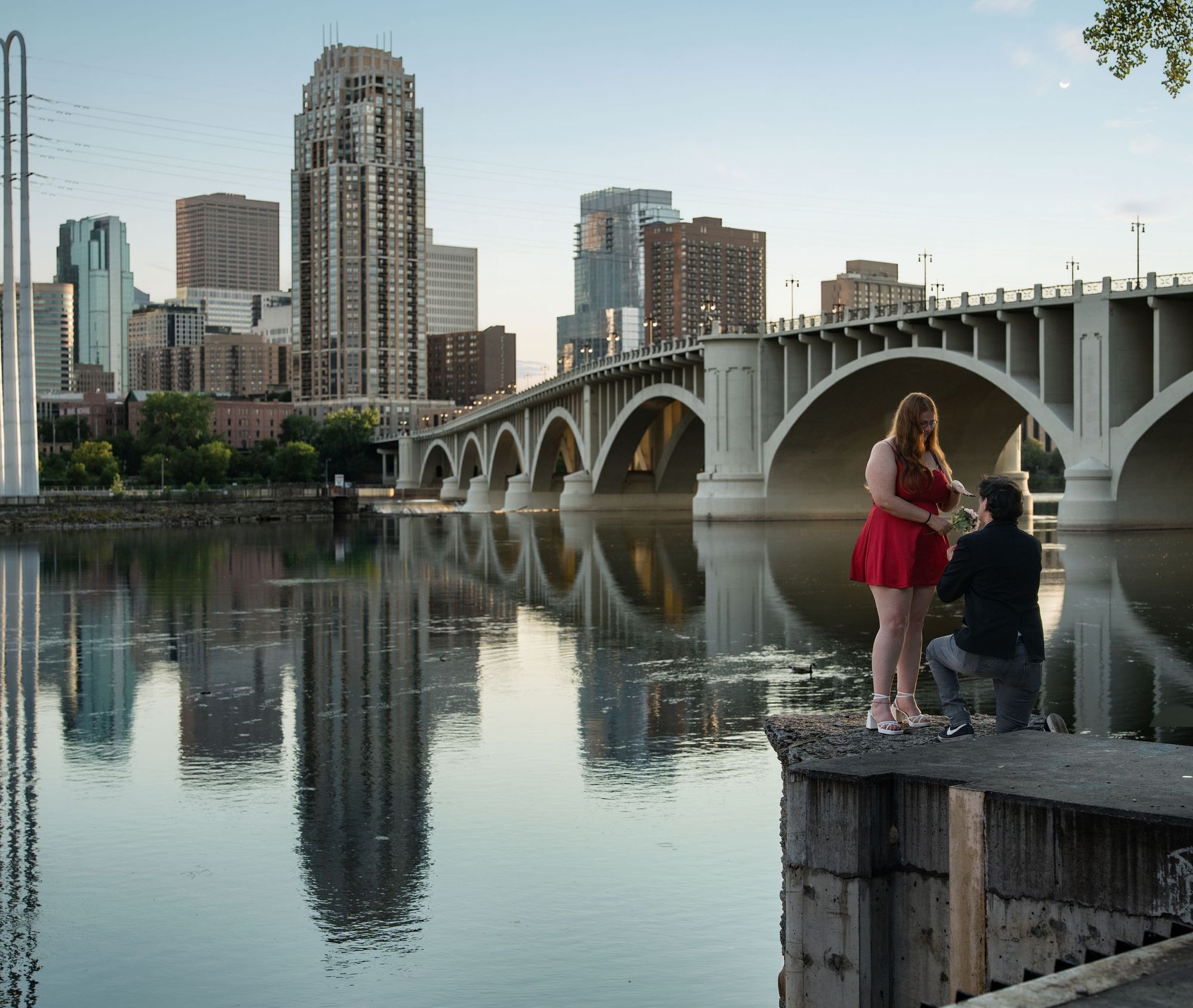 A man kneels to propose on a riverside ledge beside an arched bridge and downtown skyline