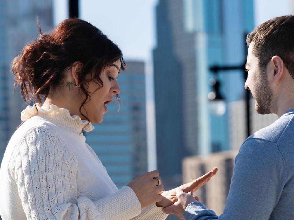 A woman admires her new engagement ring as her partner looks on with city towers behind