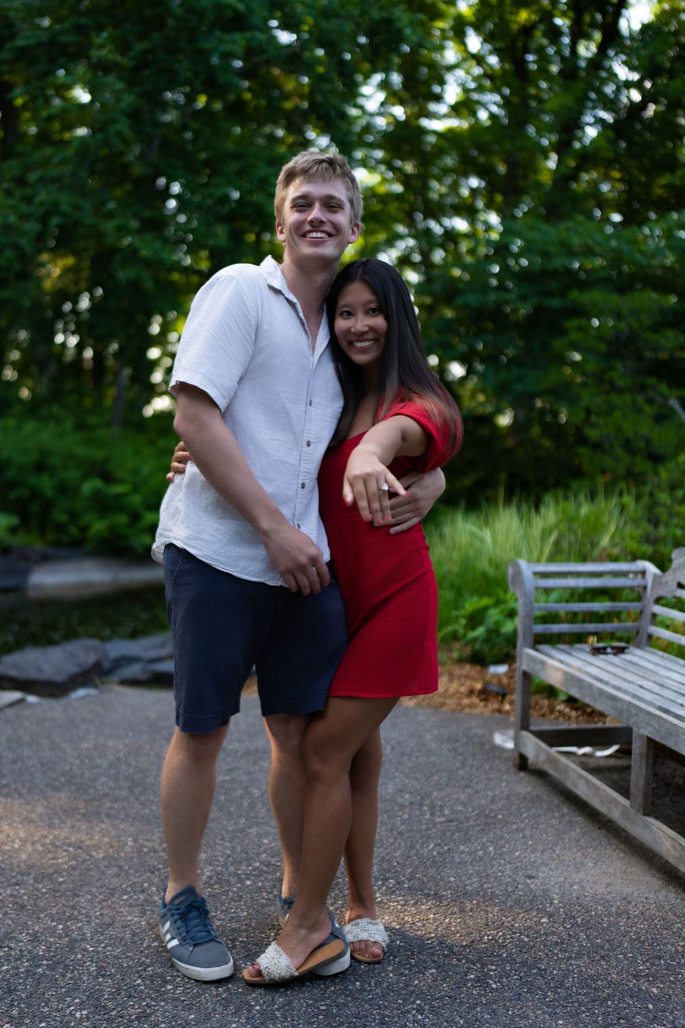 A newly engaged couple smiles as the woman shows her ring on a garden path