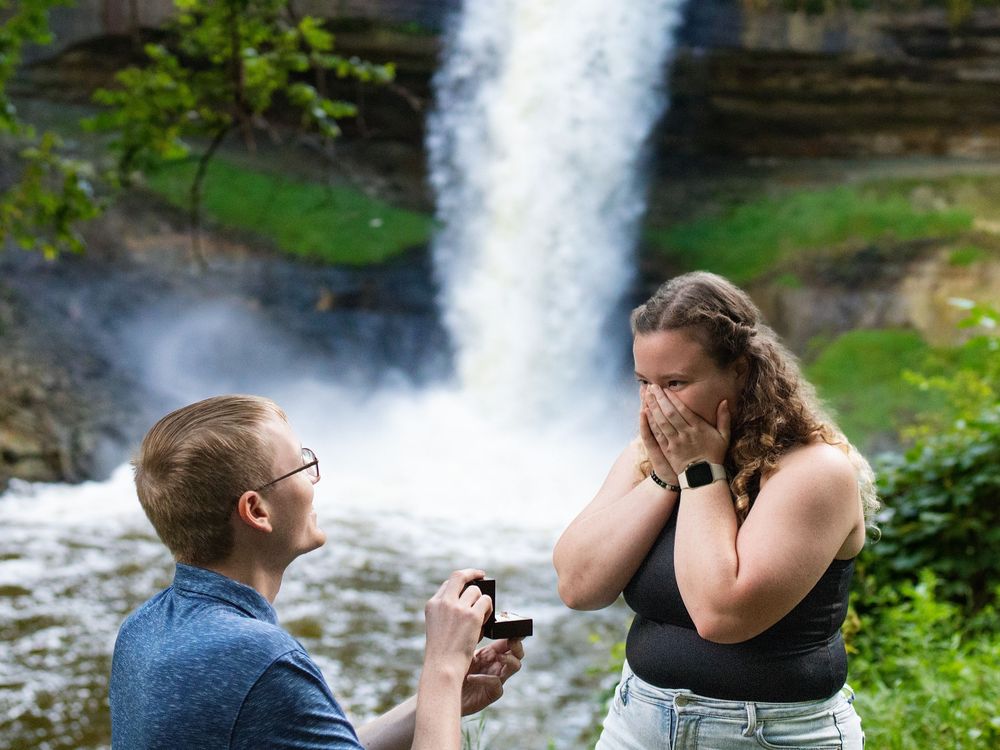 A man kneels with a ring as a woman covers her face in surprise before a waterfall