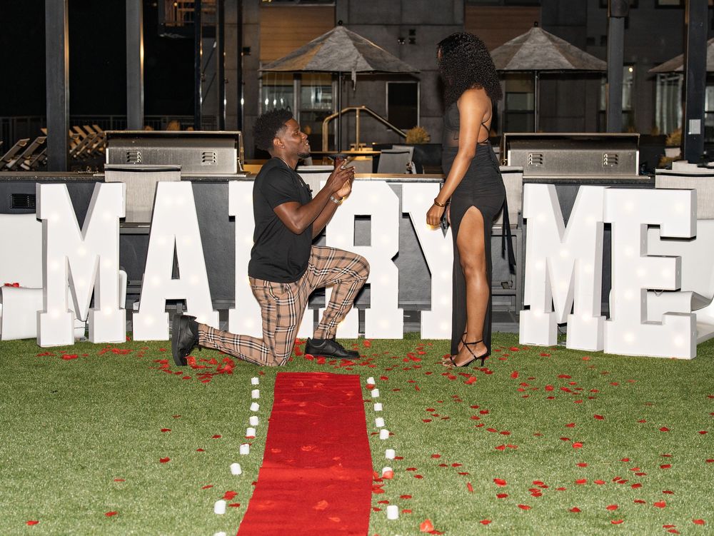 A man kneels to propose to a woman before lit MARRY ME letters and a red carpet