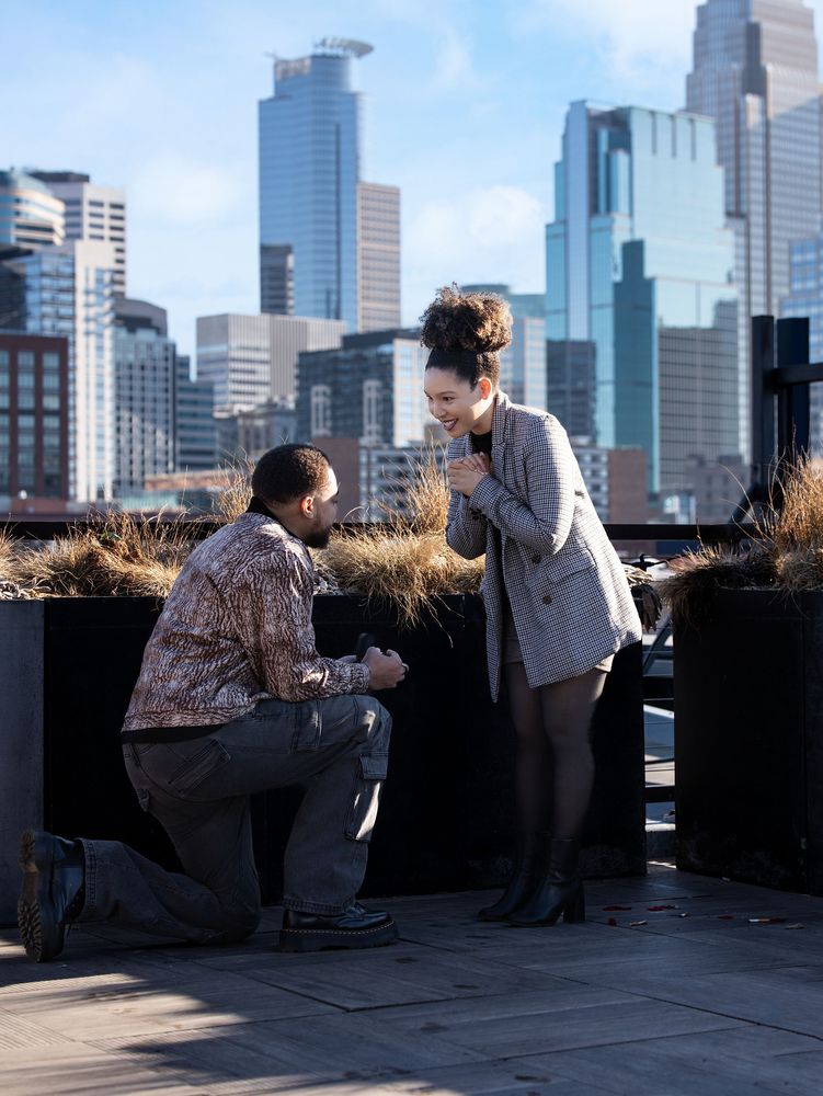A man kneels to propose to a woman on a rooftop with the Minneapolis skyline behind
