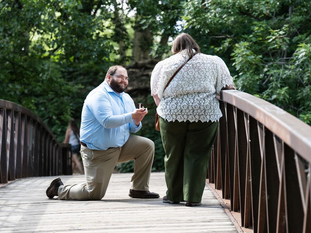 A man kneels to propose to a woman on a wooden footbridge surrounded by trees