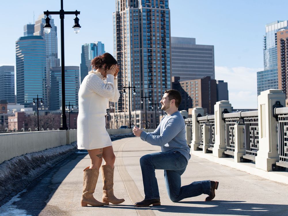 A man kneels to propose on a stone bridge as a woman covers her face, skyline behind