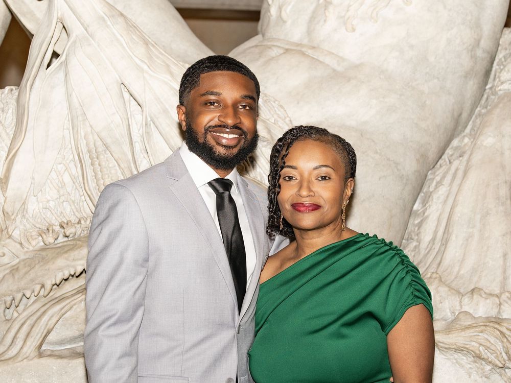 Two wedding guests, a man in grey and a woman in green, pose before a marble sculpture