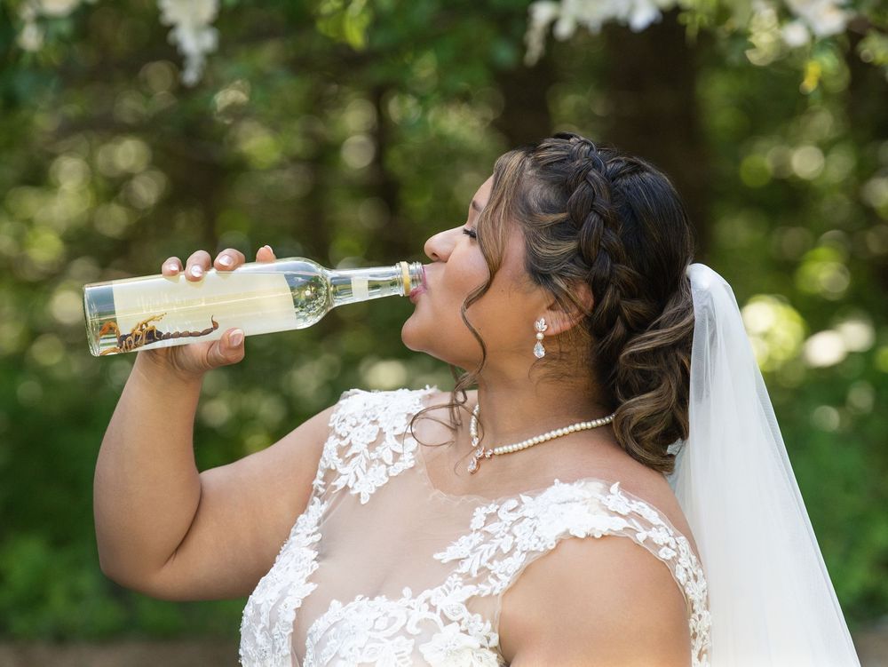 A bride in a lace gown and veil playfully drinks from a wine bottle outdoors
