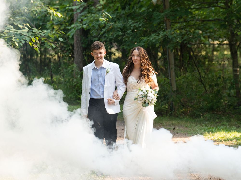 A young man in a white jacket walks the bride down a wooded aisle through white smoke