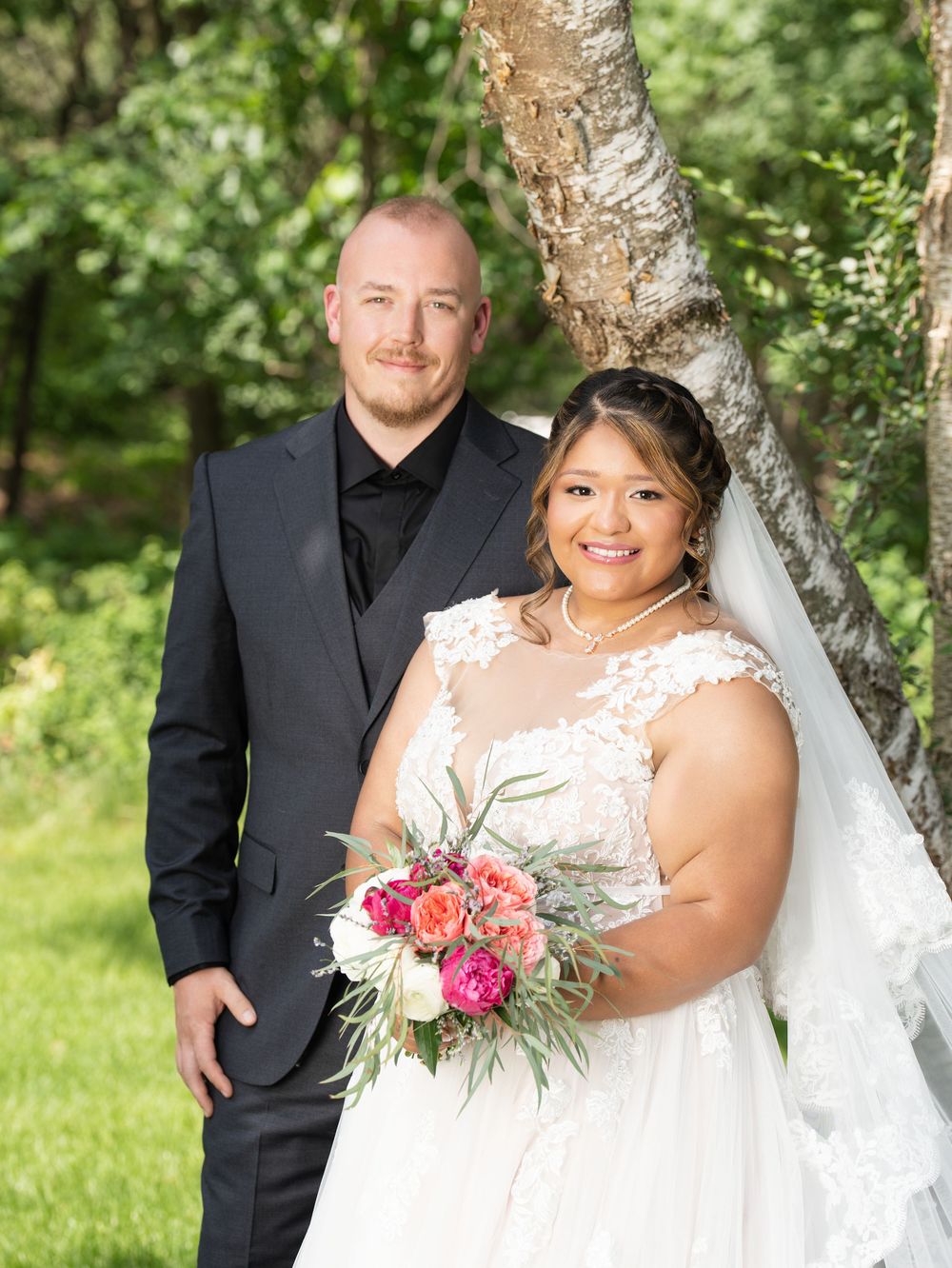 A bride holding a pink bouquet stands with the groom beside a birch tree