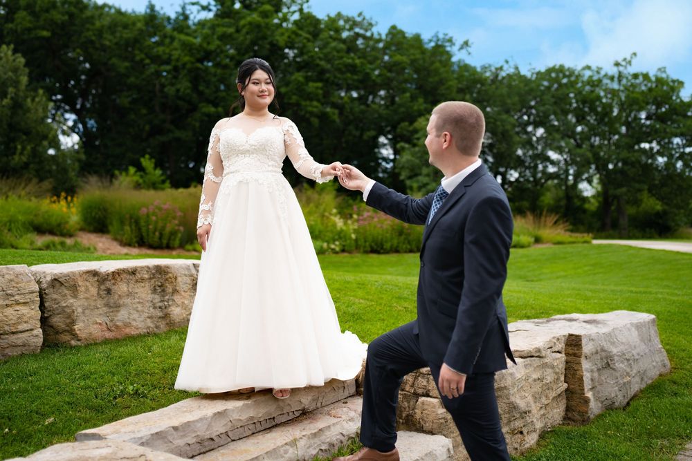 A groom holds the bride's hand as she steps along a stone wall in a garden