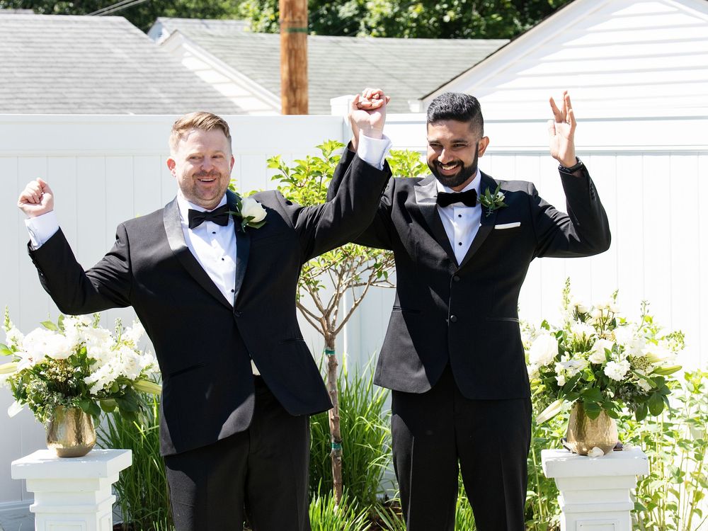 A newlywed couple in black tuxedos raise joined hands and cheer at a backyard ceremony