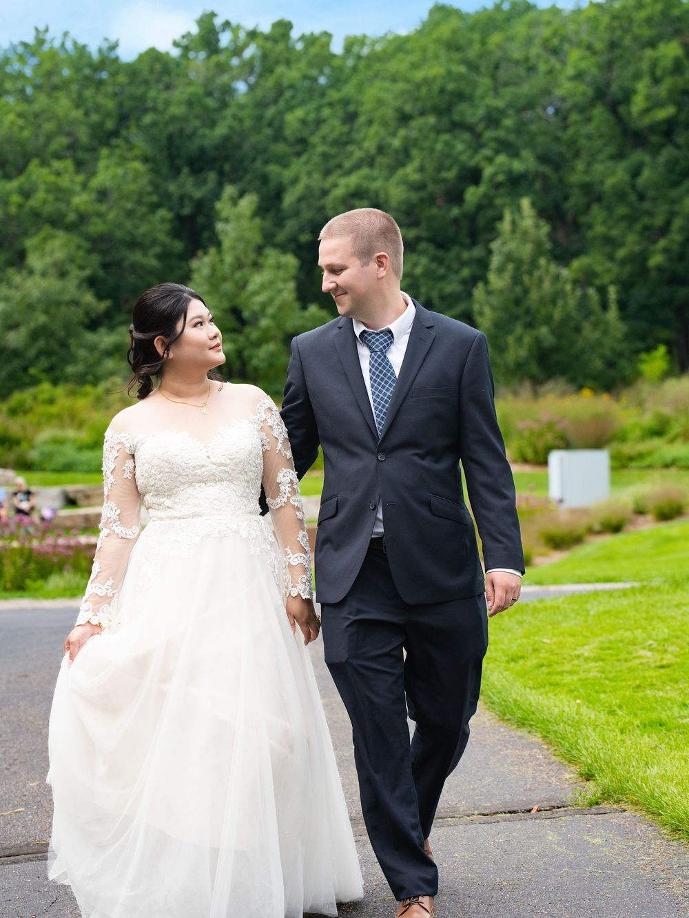 A bride and groom walk hand in hand along a garden path, smiling at each other