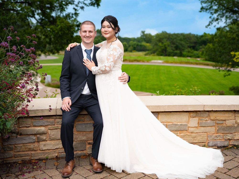 A bride and groom pose together against a low stone wall overlooking a green lawn