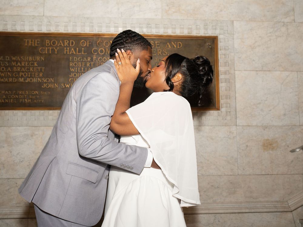 A groom in a grey suit kisses his bride beside a bronze plaque in a marble hallway
