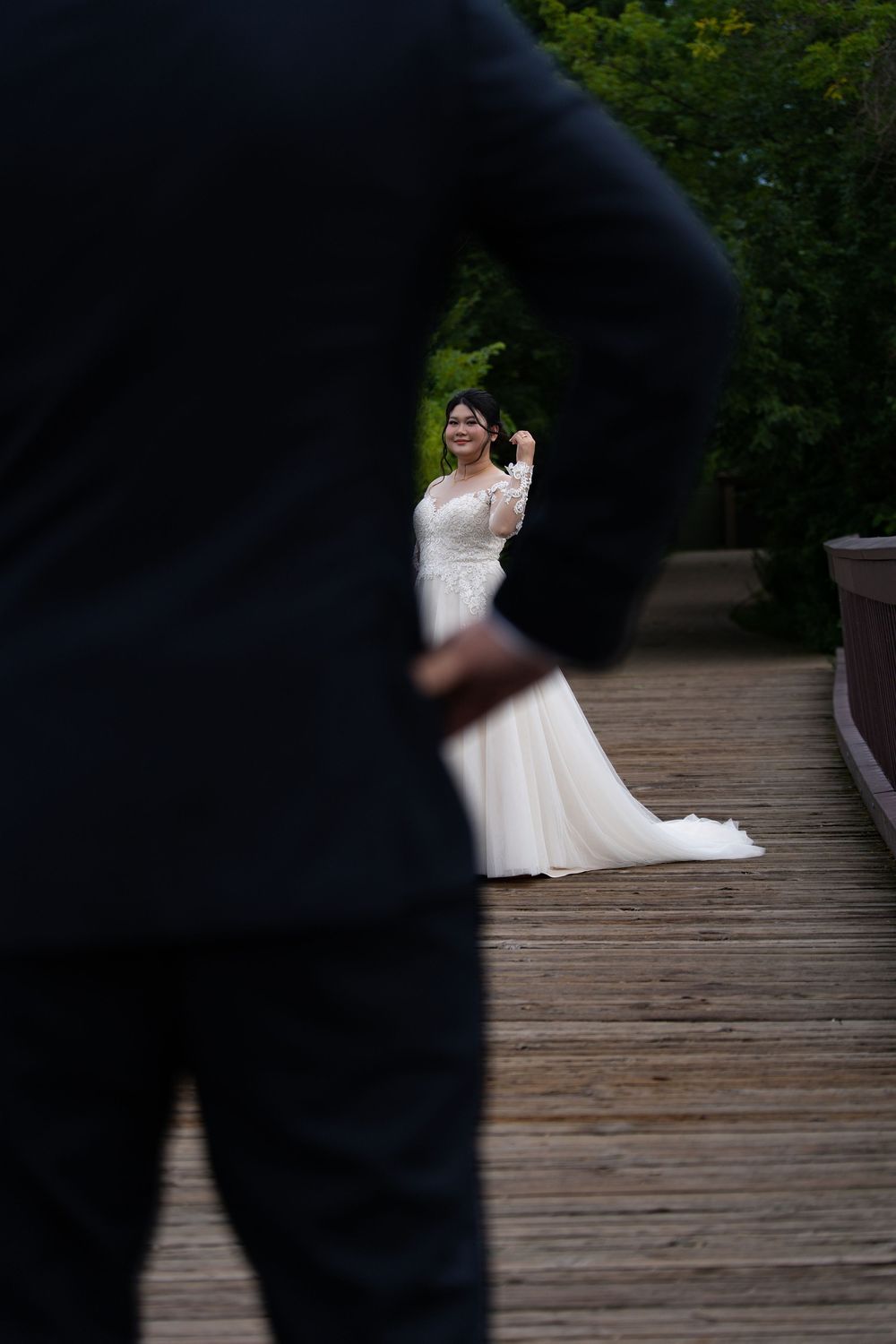 A groom in the foreground watches his bride approach on a wooden boardwalk during their first look