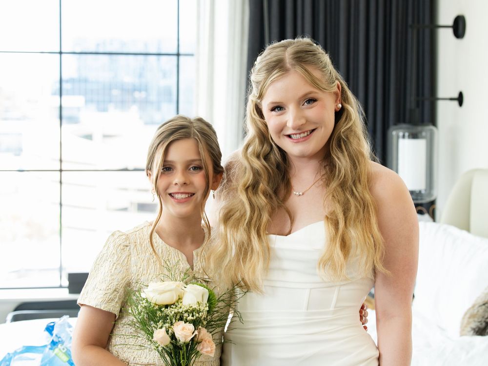 A bride in a strapless gown smiles with a young girl holding a bouquet in a hotel suite