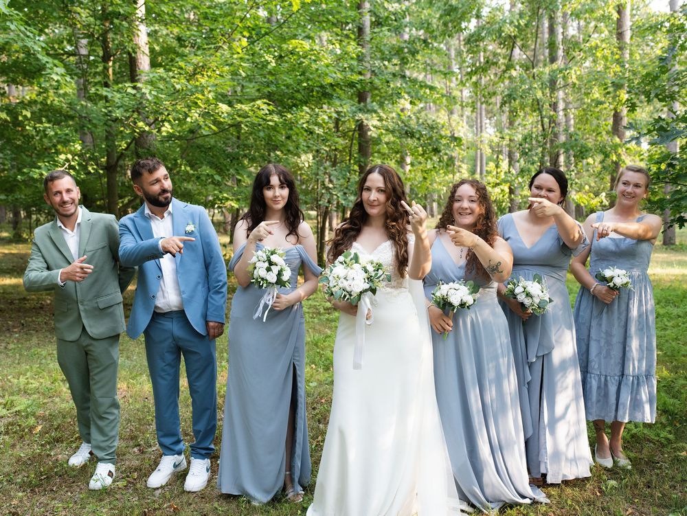 A bride shows off her ring as her wedding party points to it in a pine forest