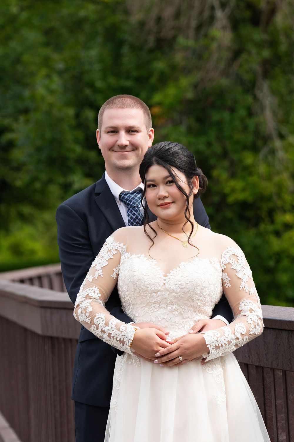A groom embraces his bride from behind on a wooden bridge surrounded by greenery