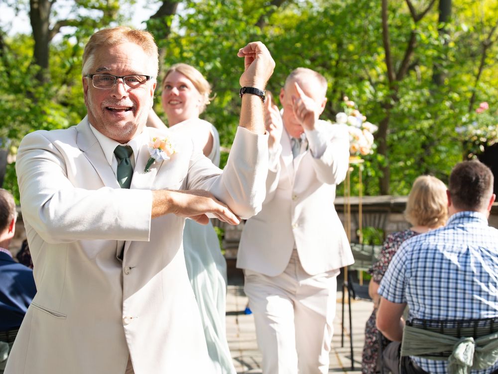 Wedding party members in cream suits dance down the aisle past seated guests at an outdoor ceremony