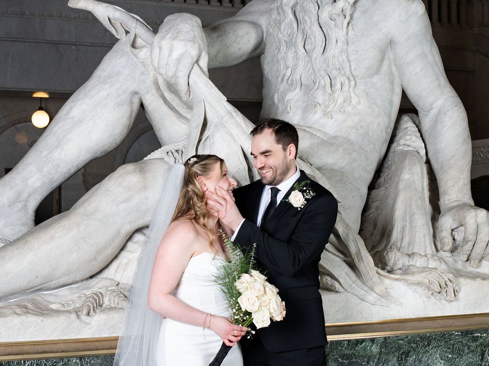 A bride holding white roses touches the groom's face beside a large marble statue
