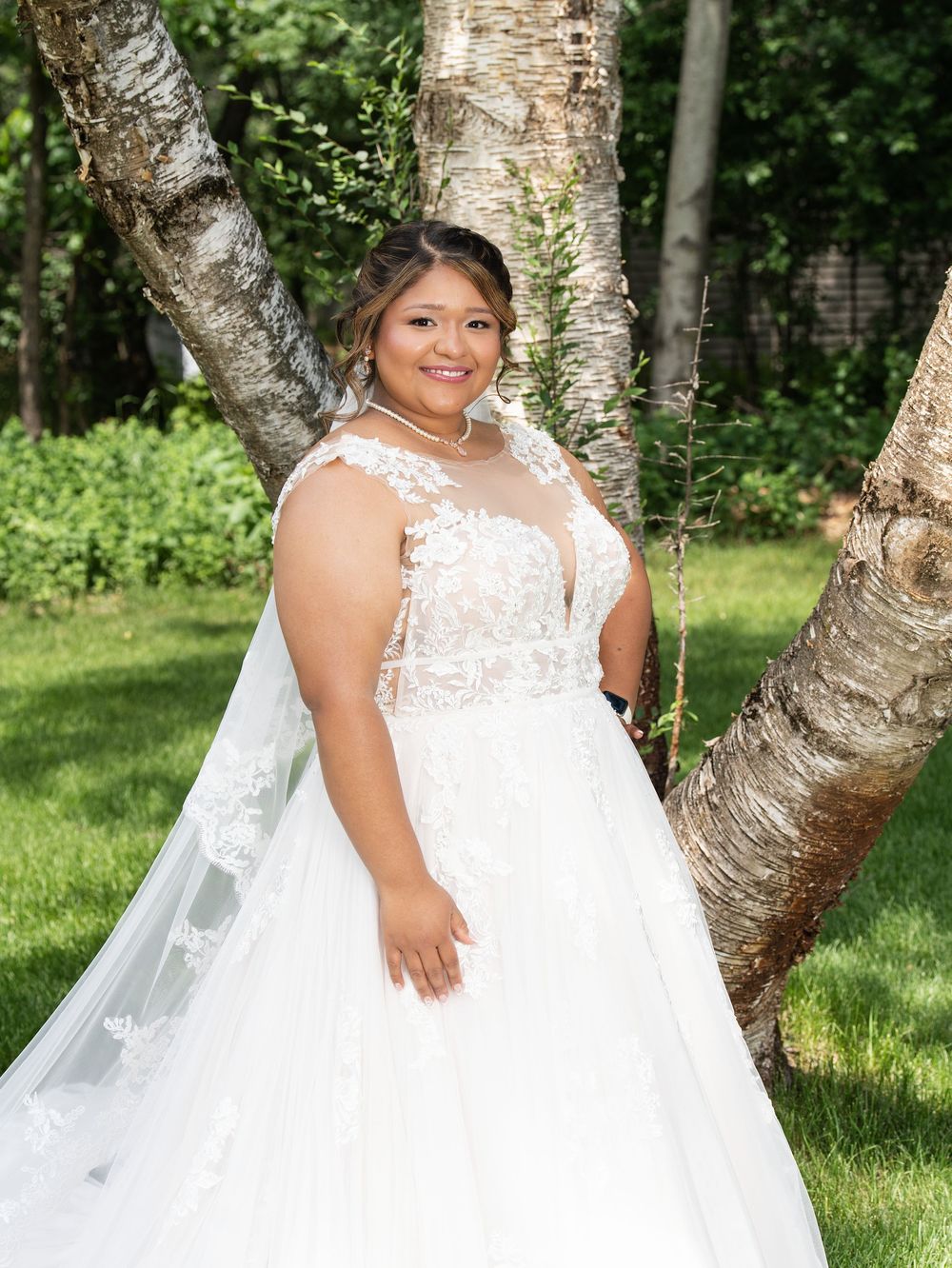 A bride in a lace ball gown and long veil stands between birch trees on a lawn