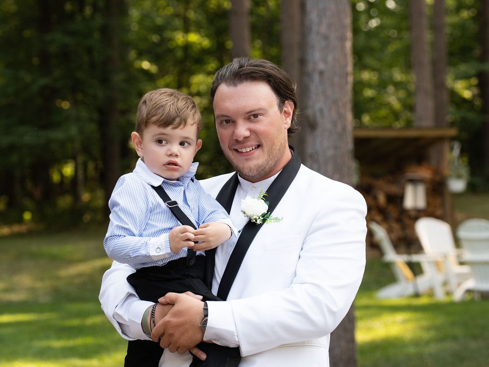 A groom in a white tuxedo jacket holds a smiling toddler on a wooded lawn
