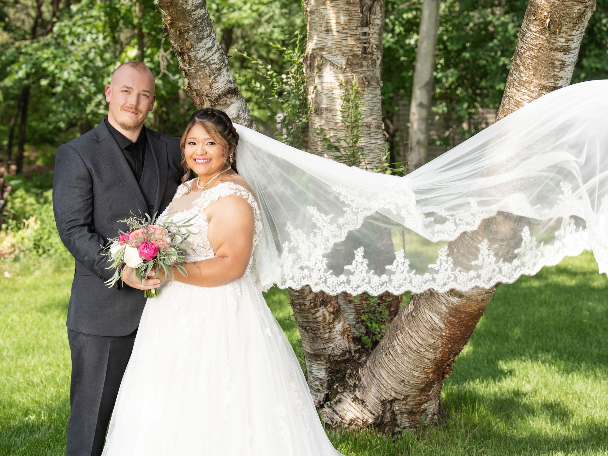 A bride's long lace veil billows in the wind as she stands with the groom under birch trees