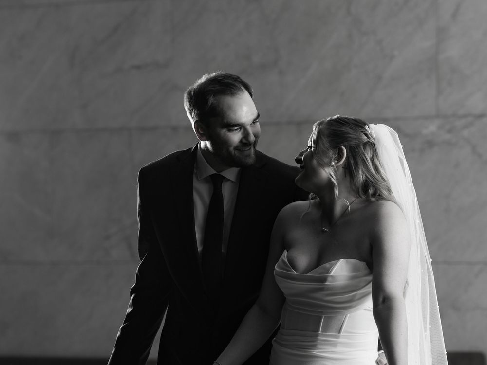 A bride and groom laugh together against a marble wall in black and white